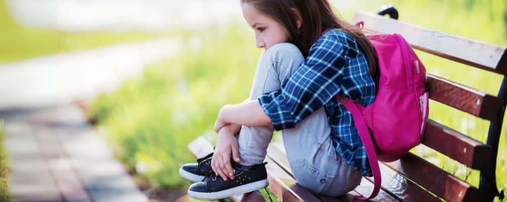 Przemoc domowa i przemoc psychiczna wobec dzieci &ndash; jak ją rozpoznać i przerwać? - smutna dziewczynka będąca ofiarą przemocy w szkole siedzie z plecakiem na ławce - blog Marita Woźny